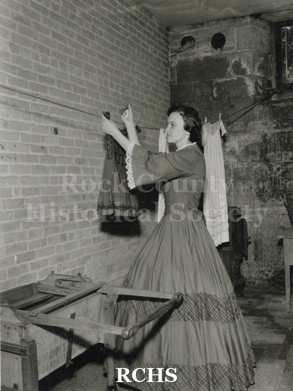 Black and white photograph of Gwendolyn C. Hughes in period dress hanging up clothes on a line in the basement of the Tallman House laundry room.