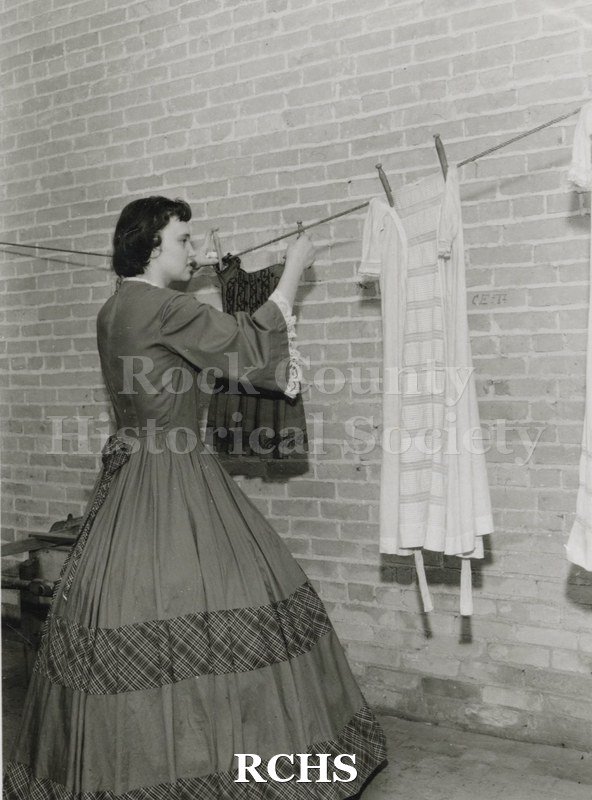 Black and white photograph of Gwendolyn C. Hughes in period dress hanging up clothes on a line in the basement of the Tallman House laundry room.
