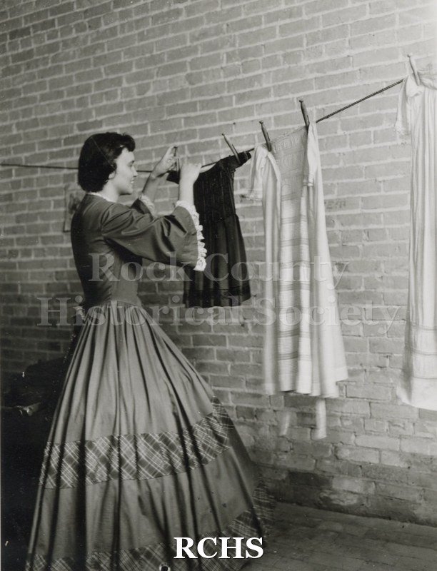 Black and white photograph of Gwendolyn C. Hughes in period dress hanging up clothes on a line in the basement of the Tallman House laundry room.