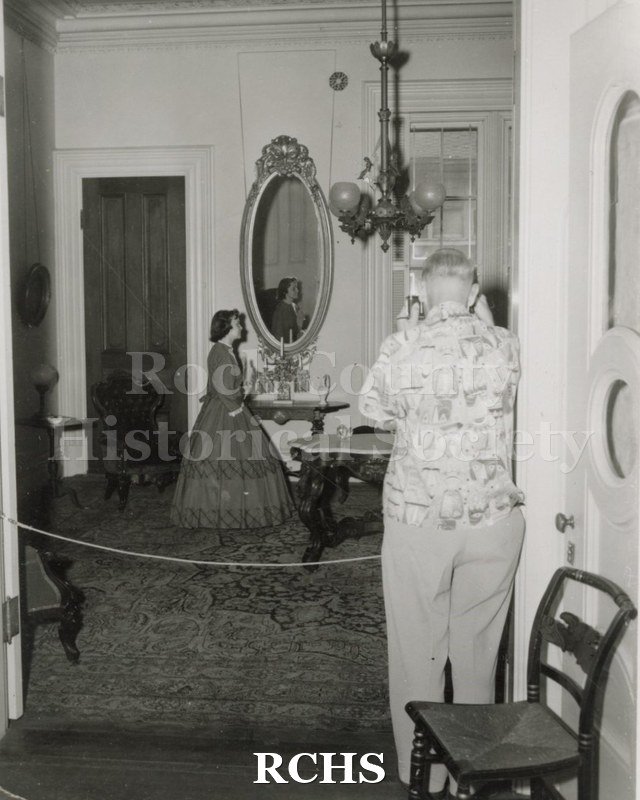 Black and white photograph of Gwendolyn C. Hughes in period dress standing near a table in the Tallman House.
