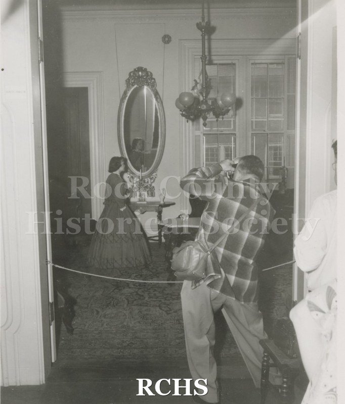 Black and white photograph of Gwendolyn C. Hughes in period dress standing near a table in the Tallman House.