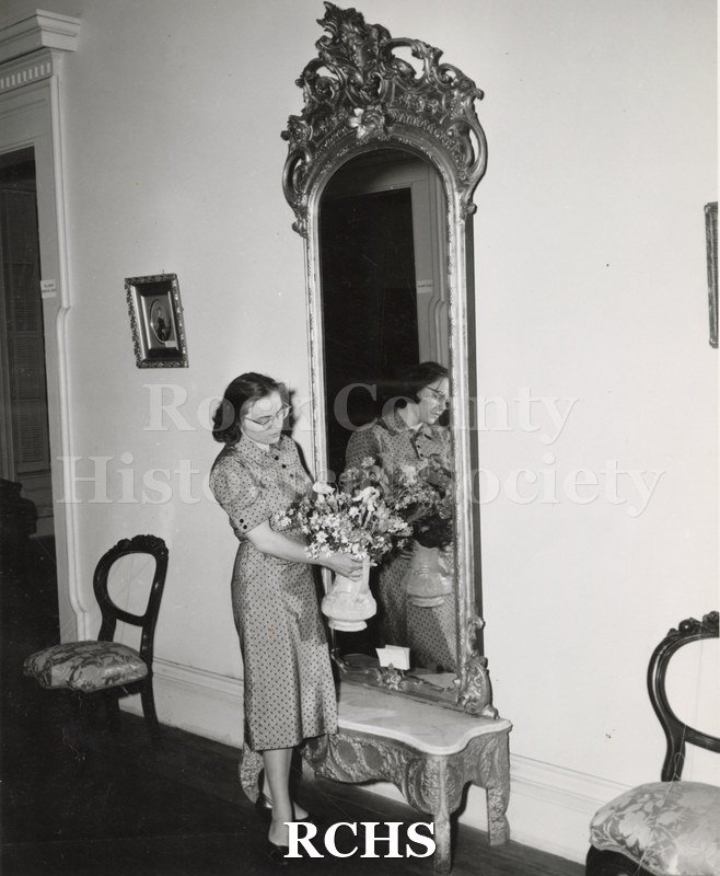 Black and white photograph of Mrs. Robert Hall holding a vase of flowers in the Tallman House. She is about to set it in front of a mirror with a marble base.