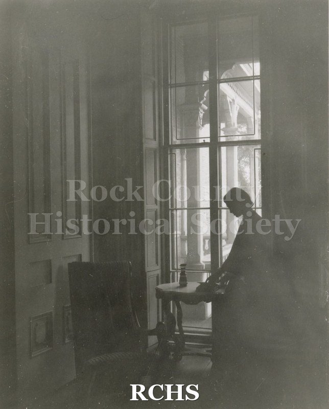 Black and white photograph of Gwendolyn C. Hughes in period dress in silhouette standing near a table in front of the window at the Tallman House.