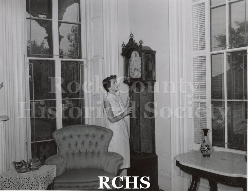 Black and white photograph of an unidentified young woman standing in front of the grandfather clock in the corner of the parlor in the Tallman House.