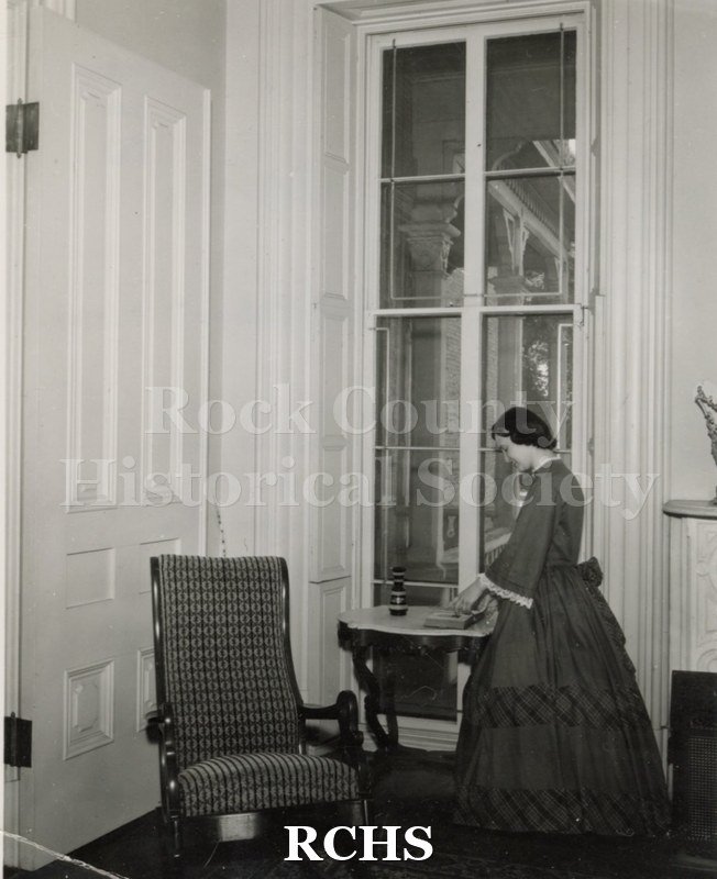 Black and white photograph of Gwendolyn C. Hughes in period dress standing near a table in the Tallman House. She appears to be looking at a book.
