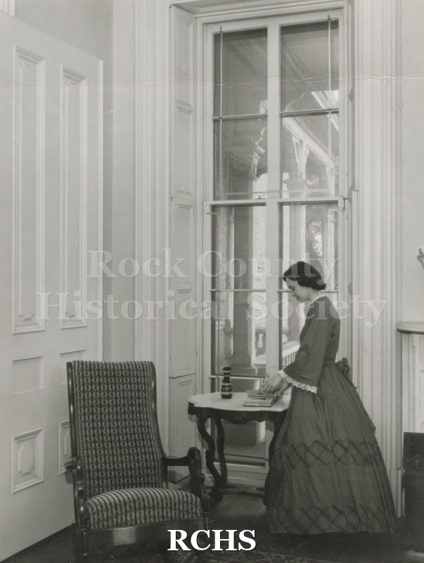 Black and white photograph of Gwendolyn C. Hughes in period dress standing near a table in the Tallman House. She appears to be looking at a book.