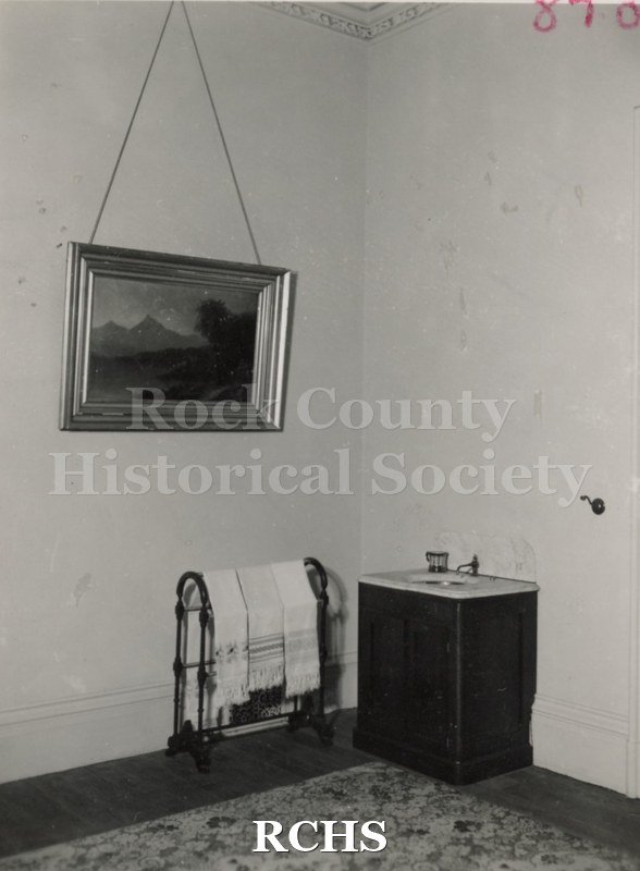 Black and white photograph of part of a bedroom in the Tallman House, showing a towel rack with towels beside an enclosed basin with one faucet.