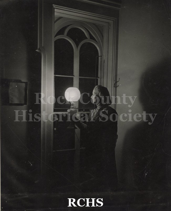 Black and white photograph of Helen Hook standing in a darkened room holding a lighted lantern lamp. She is standing beside an arched window in the Tallman House.