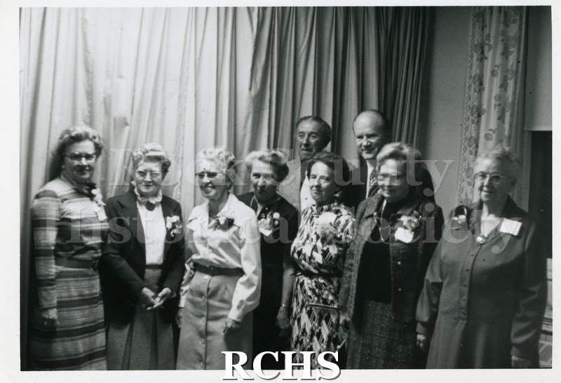 Historic image of Charter Members United Methodist Church, Evansville, WI. Shows several women and men standing in front of draperies. Women are wearing corsages.