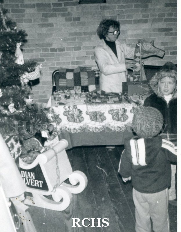 Black and white photograph of Esther Hodge standing beside a craft table. There are children standing in front of the table.