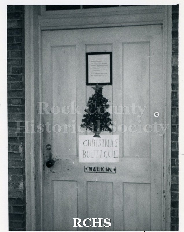 Black and white photograph of the outside of the Carriage House door with a sign "Christmas Boutique" and a small tree decoration on it.