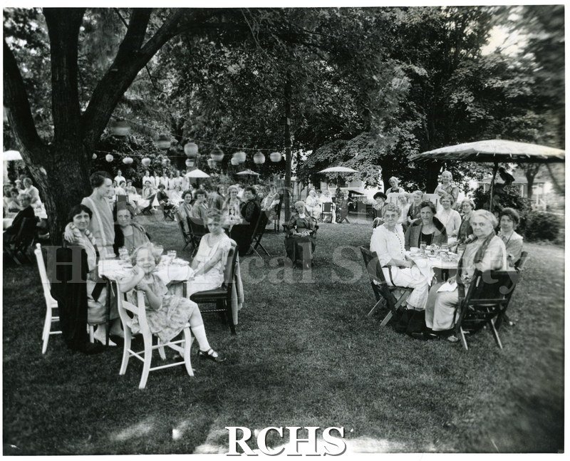 Black and white photograph of a garden party in Edgerton. Shows a group of people at a picnic. They are seated at tables on the lawn. Some tables have umbrellas.