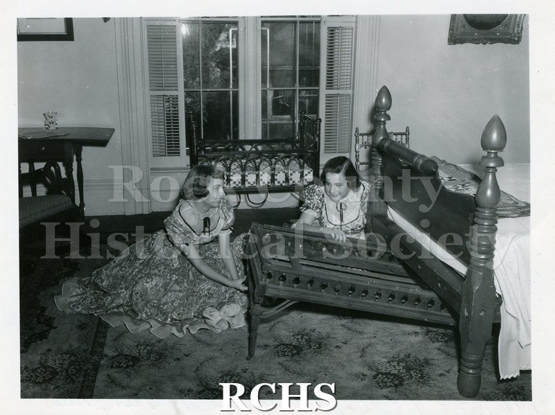 Historic image of Two unidentified young women dressed in period costumes are removing the trundle bed from under the main bed in a bedroom of the Tallman House.