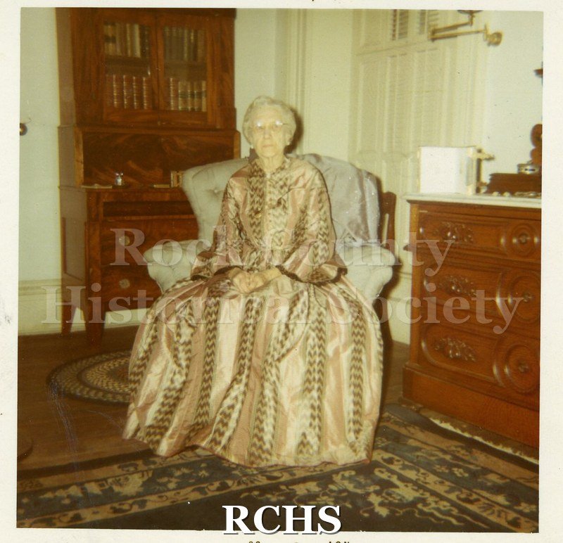 Black and white photograph of Mrs. Effie Adams wearing a full skirted long sleeved dress while sitting in an upholstered chair in a room in the Tallman House.