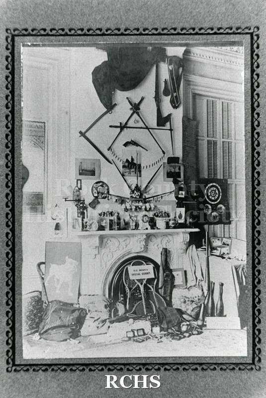 Black and white photograph of the Tallman Gun Collection. Shows a fireplace with guns and swords mounted on the wall above it. There are various items on the mantel.