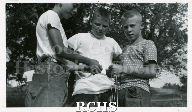 Black and white photograph of three boys working on some string.