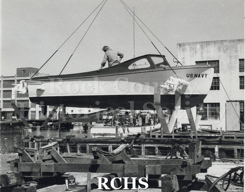 Black and white photograph of "High Pockets" on a hoist above the water. There is a man in the boat looking down. The sign "U.S.Navy" is visible.