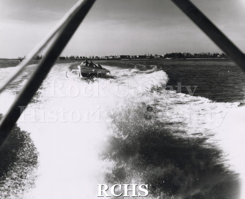 Black and white photograph of "High Pockets" in water at Patuxent Naval Air Station, MD, going through the wake of another boat. There are three men aboard.