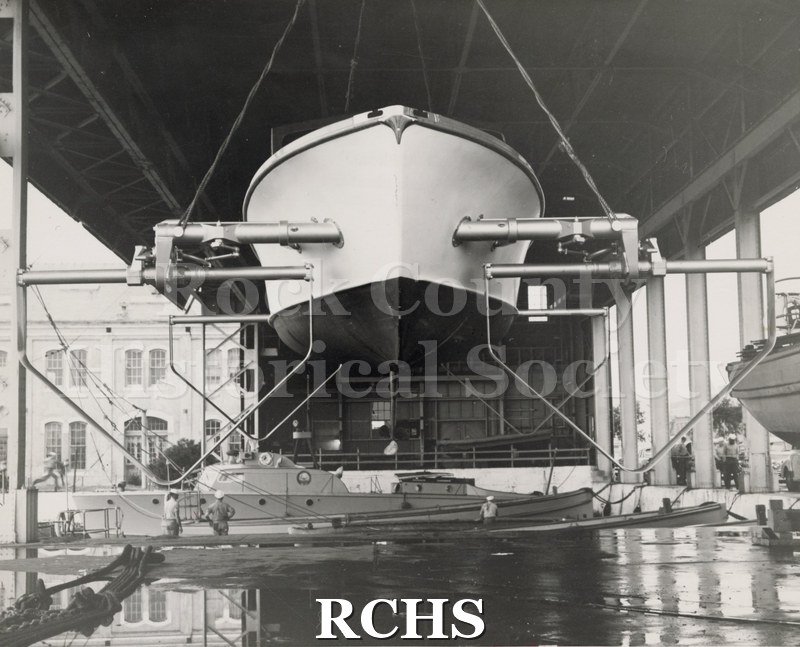 Black and white photograph of "High Pockets" on a hoist at naval yard with another naval vessel below it in the water. There are sailors standing along the way.