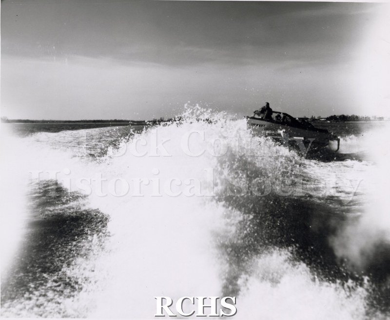 Black and white photograph of "High Pockets" in the water with three men aboard. There is a wide wake in front of it.
