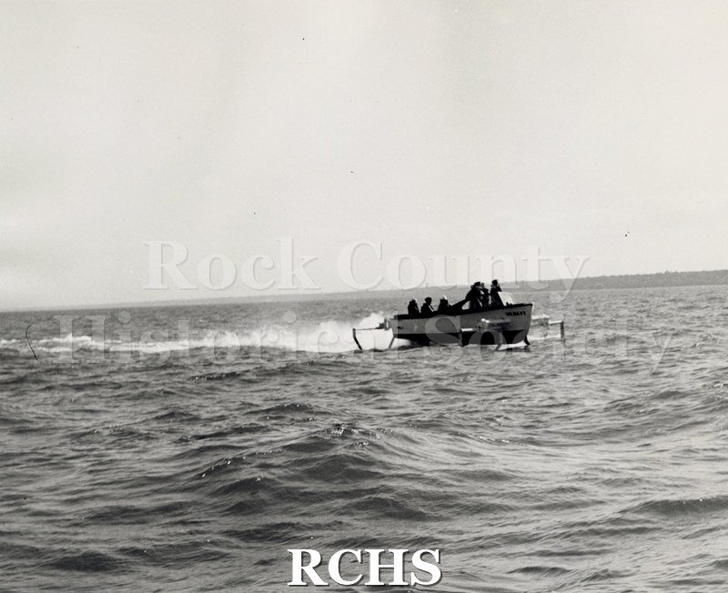 Black and white photograph of "High Pockets" in the water with several men aboard. There is a small wake behind the boat.