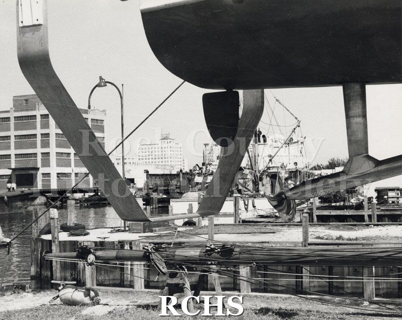 Black and white photograph of "High Pockets" showing pontoon parts high on a hoist above the water.