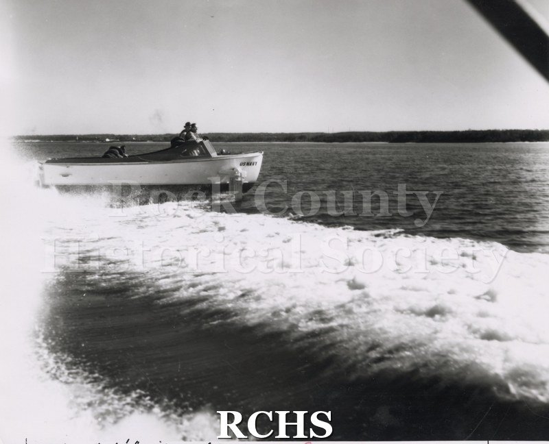 Black and white photograph of "High Pockets" on the water with three men aboard. A large wake is visible in front of the photo.