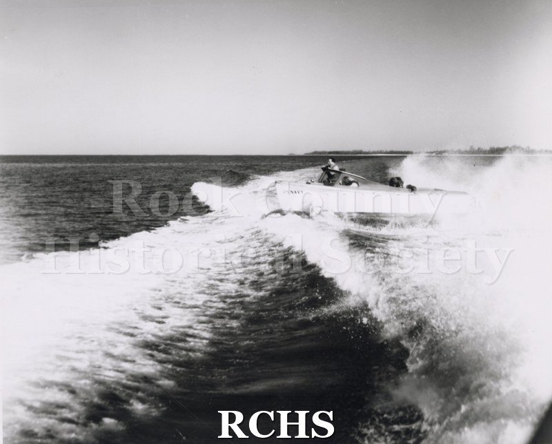Black and white photograph of "High Pockets" on the water with one man aboard. A large wake is visible in front of the photo.