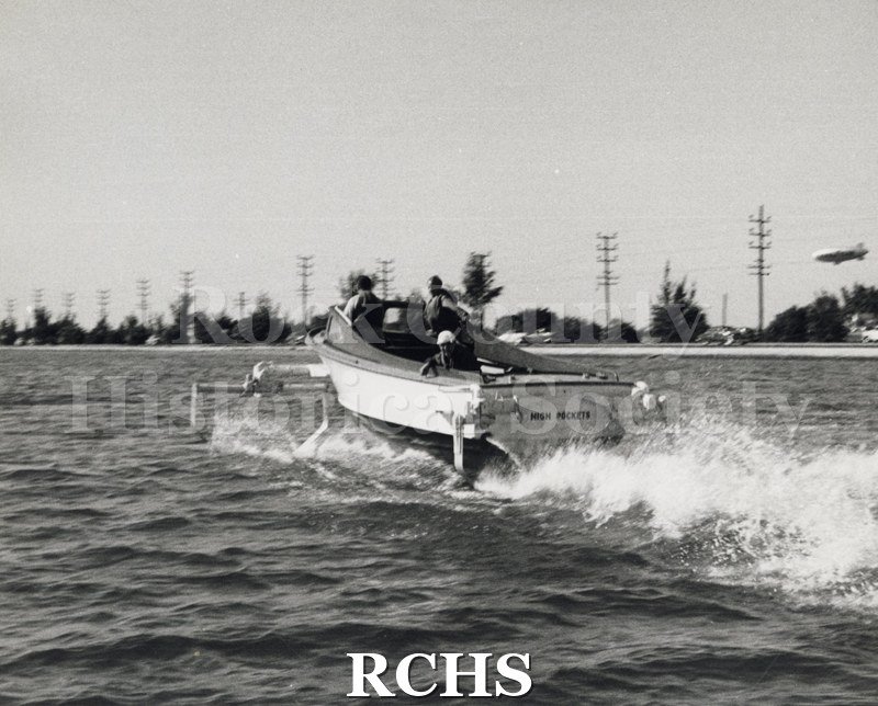 Black and white photograph of "High Pockets" with three men aboard. The name "High Pockets" is visible on the stern. A small wake is visible behind the boat.