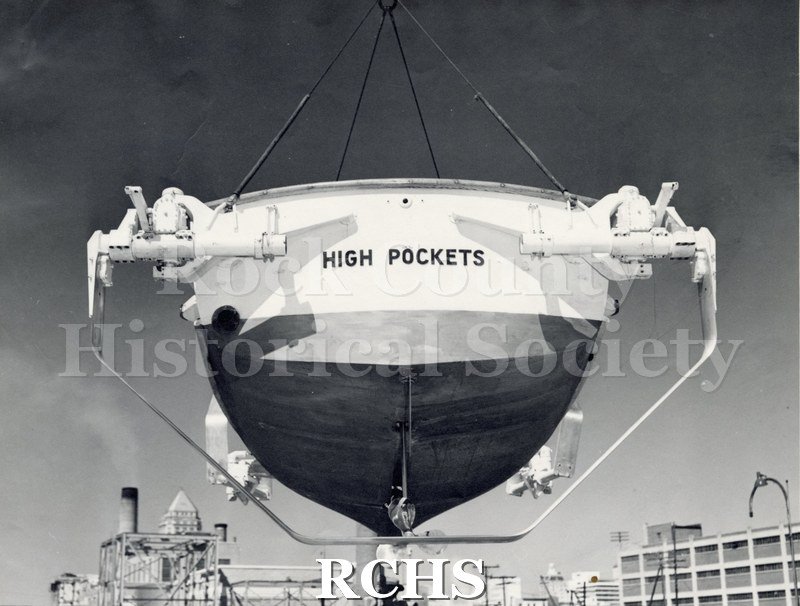 Black and white photograph of "High Pockets" on a hoist in the naval yard. Shows the back end of the boat with the words: "High Pockets" painted on it.