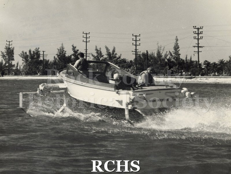 Black and white photograph of "High Pockets" with three men aboard. The boat is moving away from the camera with foils down.