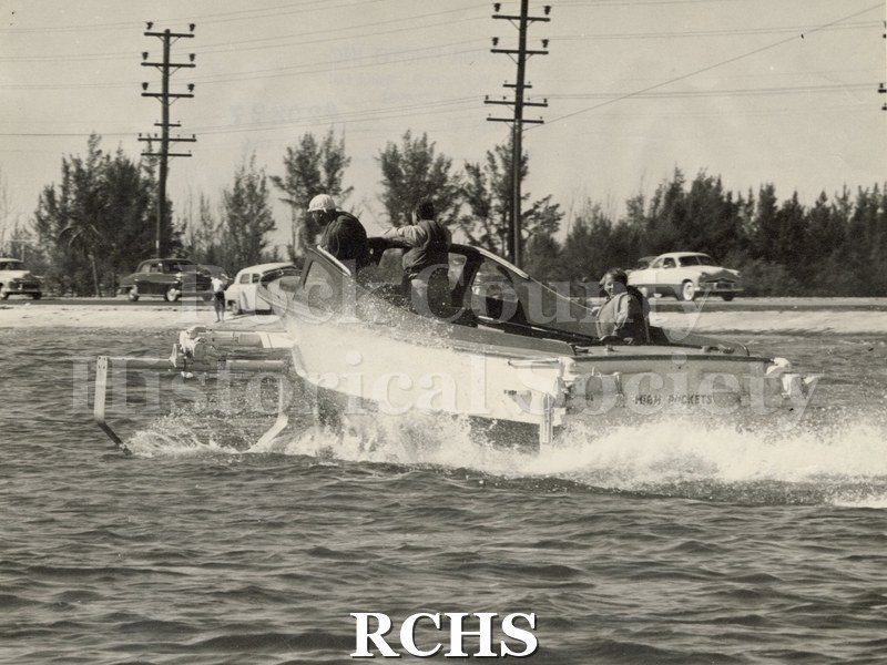 Black and white photograph of "High Pockets" with three men aboard. The boat is moving away from the camera and is near the road. Cars are visible on the road.