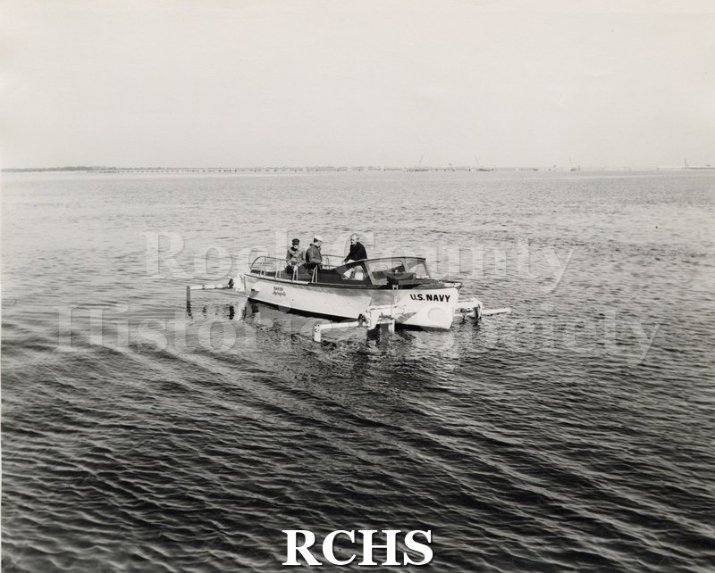 Black and white photograph of "High Tail" on the water with three men aboard. The words "U.S. Navy" are on the front and "Baker Hydrofoils" on the back.