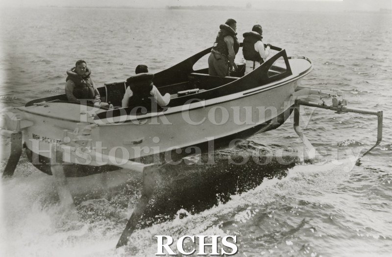 Black and white photograph of "High Pockets" on the water with four men aboard. Three are facing forward and one is looking backward.