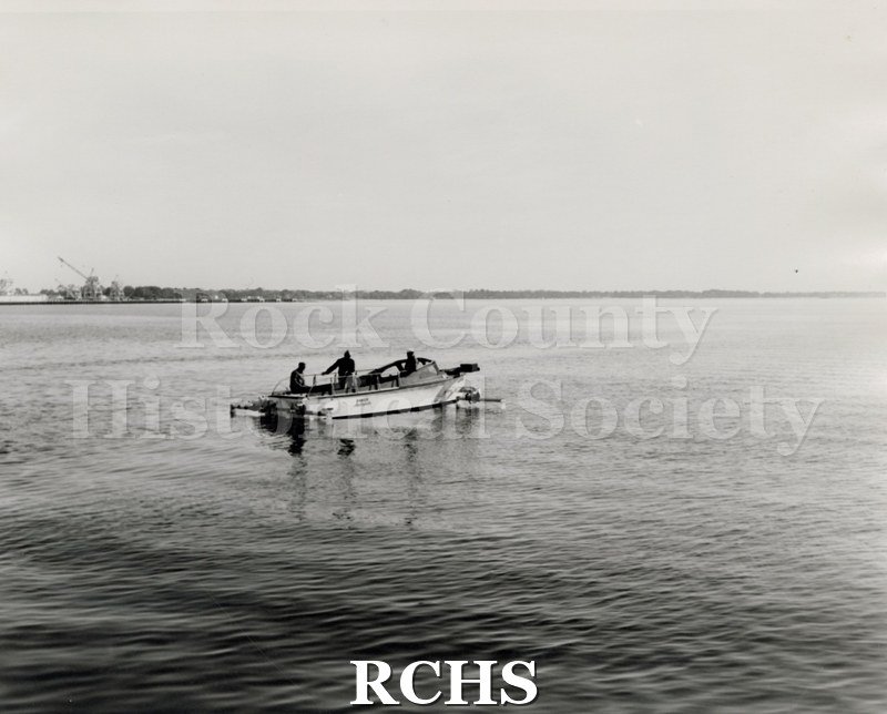 Black and white photograph of "High Pockets" on the water with three men aboard.