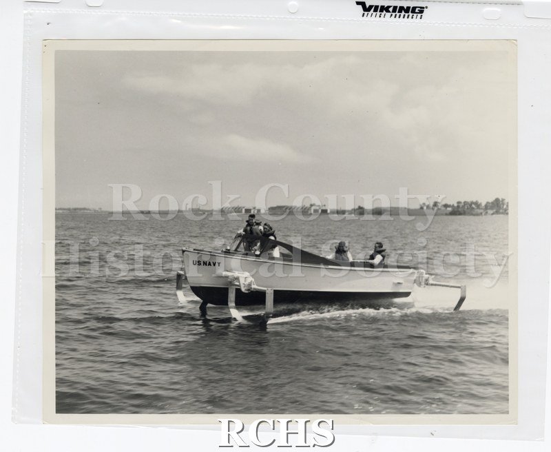Black and white photograph of "High Pockets" on the water with four men on board.. Two are standing and two are sitting in the back.