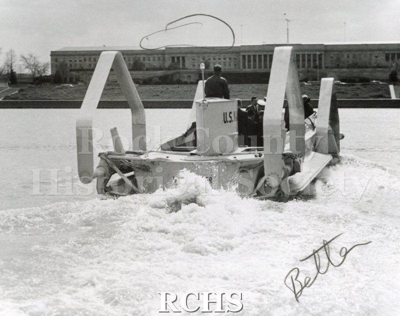 Black and white photograph of the stern of the hydrofoil with several people aboard. It is moving toward a building on the shore. On photo is the word: "Better".