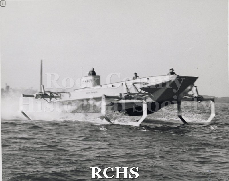 Black and white photograph of LCVP in operation with three people aboard.. "U.S.Navy" is visible on the helm and "Baker Hydrofoils" is on the side.
