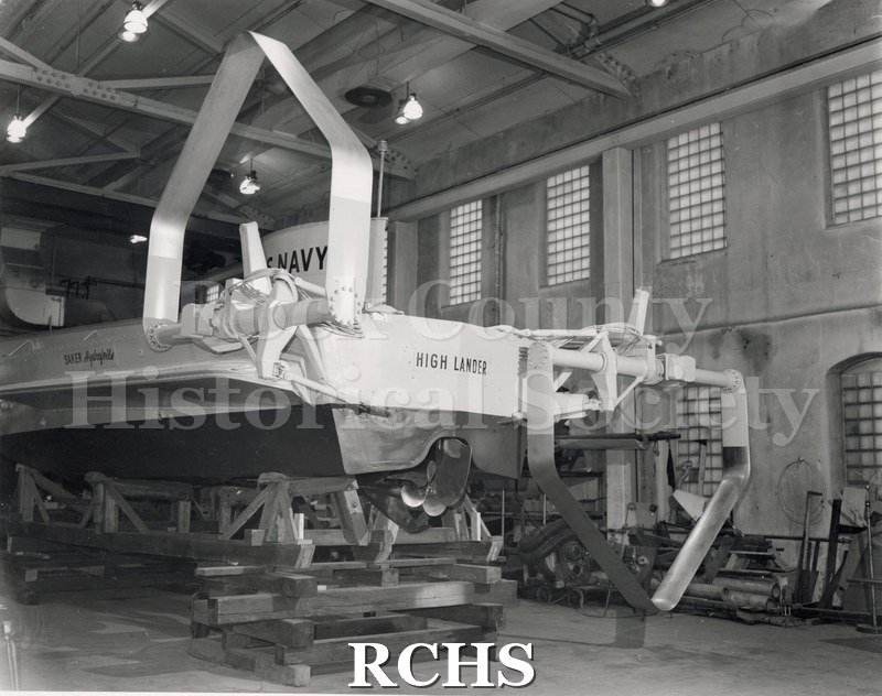 Black and white photograph of "High Lander" in the lab on a hoist. The name "High Lander" is on the back and "Baker Aquafoils" is on the side.