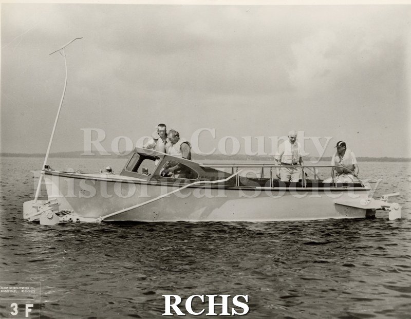 Black and white photograph of "High Tail" in the water with four men aboard. On bottom: Baker Manufacturing Co.