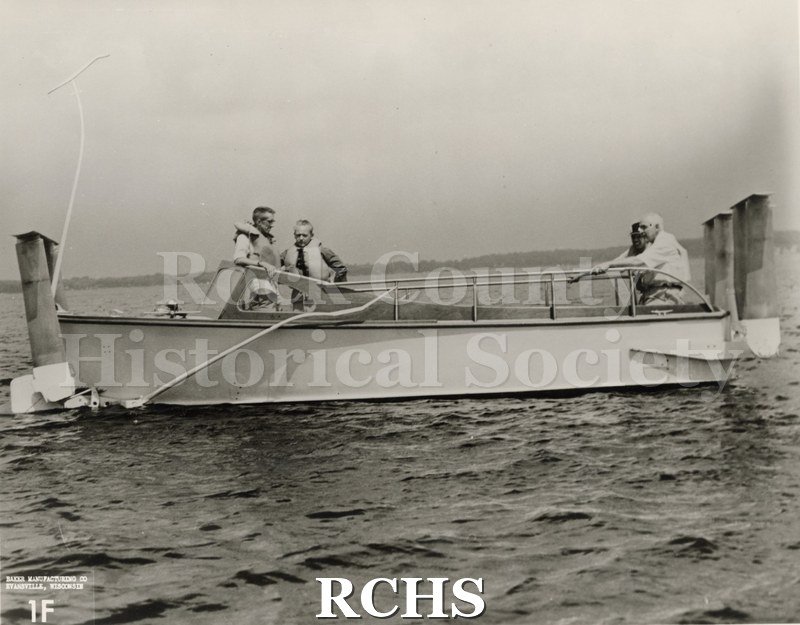 Black and white photograph of "High Tail" in the water with four men aboard. On bottom: Baker Manufacturing Co.