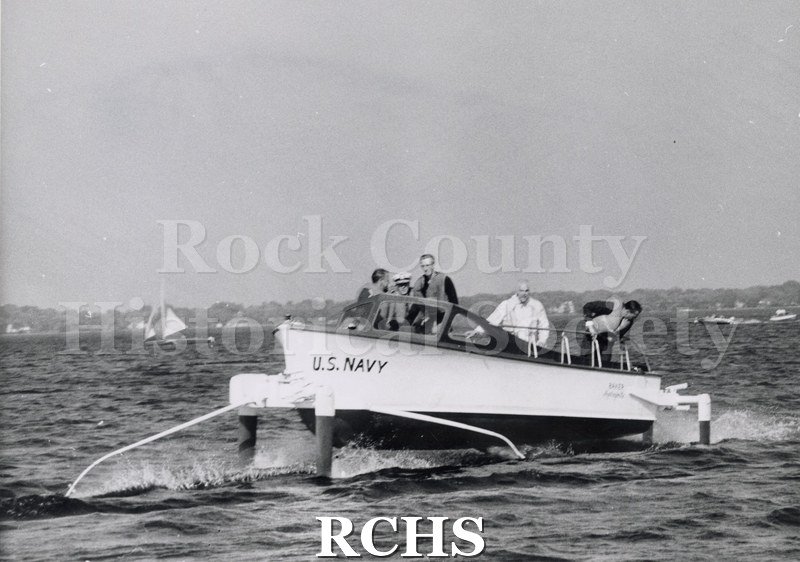 Black and white photograph of "High Tail" in the water with five men aboard. Men are facing the camera. There is a sailboat on the left side.