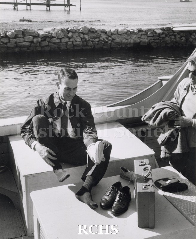 Black and white photograph of "High Tail" in the water with a man sitting in the boat changing his shoes while another man watches.