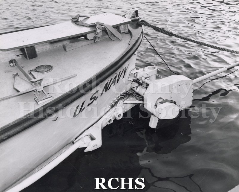Black and white photograph of a close-up shot of foil attachment-side view. Boat is in the water. Shows part of boat with "U.S.Navy" painted on it.