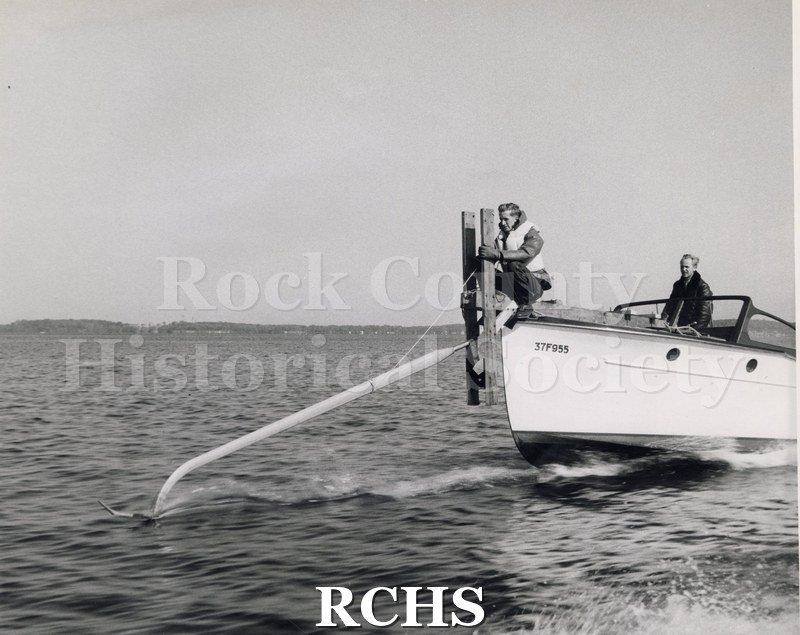 Black and white photograph of "High Tail" hydrofoil ski on the Eliza on the water with two men aboard. Shows the ski from the boat front.
