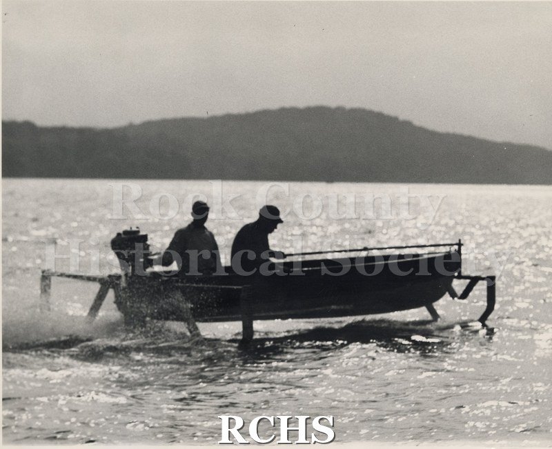 Black and white photograph of a boat with an outboard motor with two men on board.