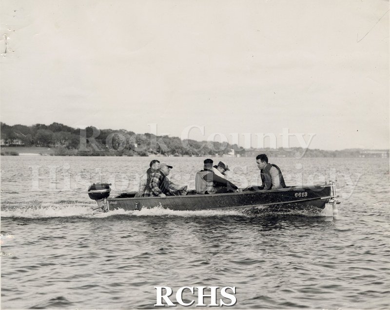 Black and white photograph of a boat with an outboard motor in the water with five men aboard. On the side is the number 6613.