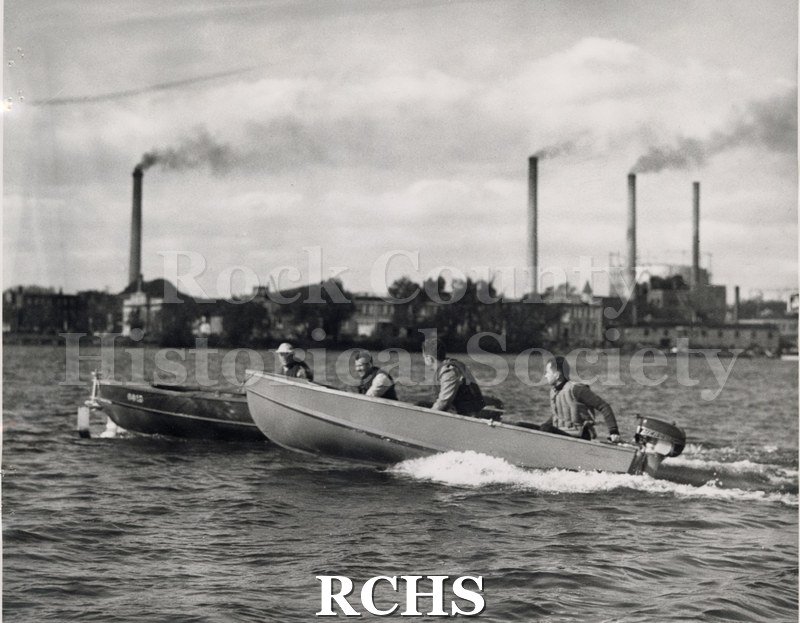 Black and white photograph of a boat with an outboard motor in the water with 3 men aboard. There is another boat beside it with one man aboard.