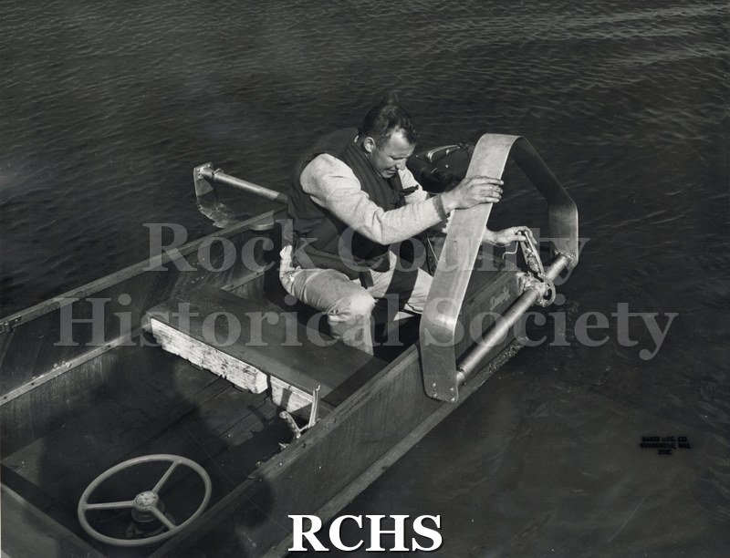 Black and white photograph of the side of a hydrofoil boat with a man adjusting the foil.