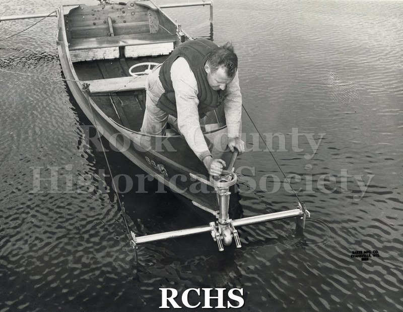 Black and white photograph of the side of a hydrofoil boat with a man adjusting the foil.